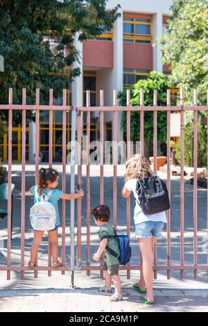 Les petites filles aux cheveux blonds et aux cheveux noirs et le petit frère aux cheveux bouclés et regardent à travers une clôture métallique fermée. Divers enfants vont à l'école. Banque D'Images