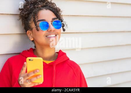 Fille adolescente jeune femme afro-américaine mixte race biracial dehors souriant avec des dents parfaites portant des lunettes de soleil bleues et en utilisant un mobi Banque D'Images