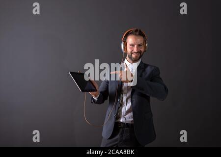 A smiling handsome young man in a suit en pointant sur l'écran d'une tablette et d'écouter de la musique devant un fond gris foncé dans un studi Banque D'Images