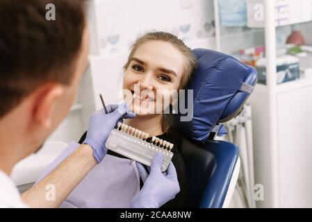 Dentiste en gants médicaux bleus appliquant l'échantillon de l'échelle d'émail des dents à la femme heureuse dents du patient pour ramasser la nuance droite, procédure de blanchiment des dents, culture, panorama. Banque D'Images