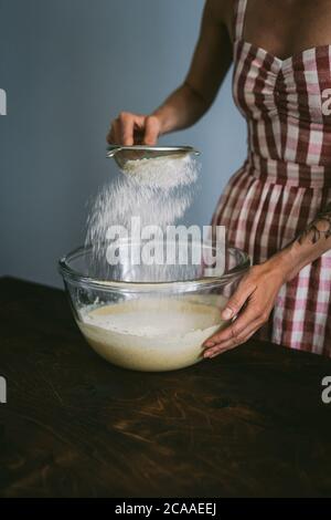 Biélorussie, Minsk - 08.03.2020: Jeune femme dans une robe à carreaux rouge et blanc qui cuit un gâteau, tamisant la farine dans un grand bol de mélange de verre avec d'autres ingredi Banque D'Images