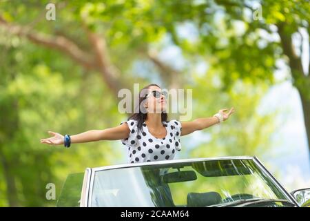 Femme en lunettes de soleil se trouve sur le dessus du cabriolet, les bras larges ouverts, se trempant le soleil et l'air frais. Banque D'Images