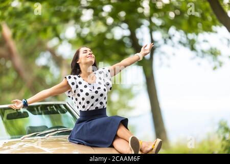 Femme en chemisier polkadot et jupe se trouve sur le capot d'une voiture cabrio, bras ouverts, profitant de l'été. Banque D'Images