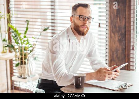 Beau homme d'affaires caucasien barbu dans un ancien t-shirt blanc, portant des lunettes, vérifiant le compte bancaire en ligne dans une tablette assise sur un canapé dans le TH Banque D'Images