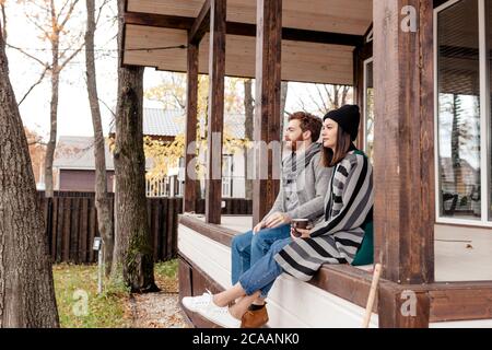 Joyeux jeune couple assis à l'extérieur, devant leur nouvelle maison en automne vêtements chauds et rêvant de leur nouvel achat passionnant. Banque D'Images