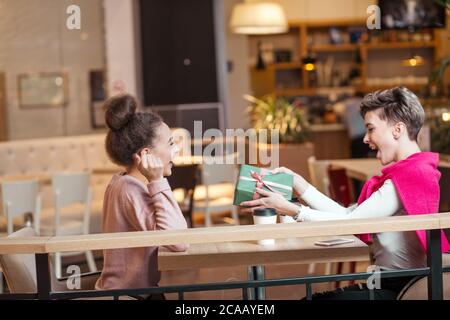 Brunette woman présente boîte-cadeau à sa copine blonde tout en restant assis dans un café du centre commercial. La fille blonde est à la recherche heureux et surpris. Shopping, les gens Banque D'Images