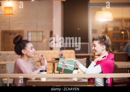 Brunette woman présente boîte-cadeau à sa copine blonde tout en restant assis dans un café du centre commercial. La fille blonde est à la recherche heureux et surpris. Shopping, les gens Banque D'Images