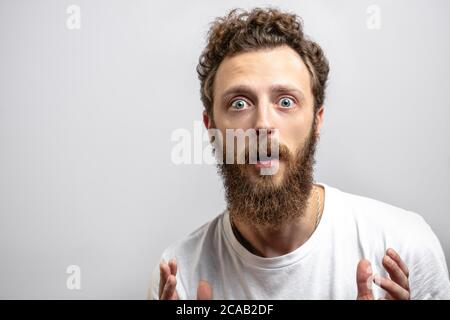 J'ai surpris l'homme caucasien avec des cheveux bouclés et des barbes avec un regard émerveillé sur l'appareil photo, vêtu d'un t-shirt blanc décontracté, isolé sur fond blanc. Banque D'Images