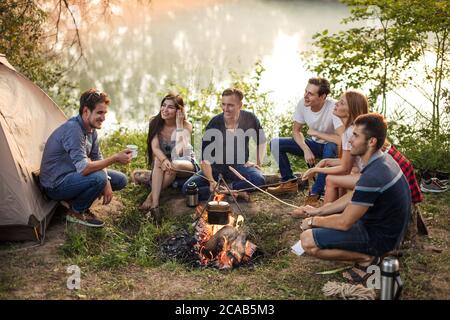 jeunes gens attrayants tenant des bâtons avec des saucisses sur le feu de camp. processus de préparation du repas dans la nature. les jeunes sont en train de se promener autour du sapin de camp Banque D'Images