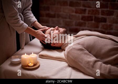 Salon de beauté, concept de traitement. Jeune homme caucasien obtenir un massage sur la tête et le visage allongé sur le bureau avec les yeux fermés, calme Banque D'Images