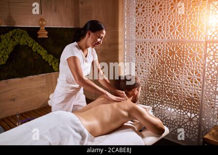 Charmante femme agréable vêtue d'un uniforme blanc, travaillant dans un hôtel spa, massant le dos d'un client masculin dans une chambre lumineuse de l'hôtel, détente totale Banque D'Images