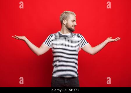 Un homme blond caucasien avec une barbe comparant deux choses dans ses paumes pose sur un mur rouge de studio Banque D'Images