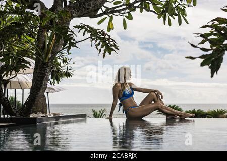 Asie, Indonésie, Bali, jeune belle femme caucasienne, portant le bikini bleu, assis sur le bord de la piscine à débordement, avec des plantes tropicales et avec la mer i Banque D'Images