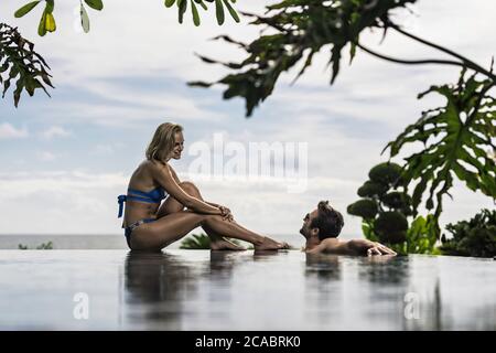 Asie, Indonésie, Bali, jeune beau couple caucasien assis au bord de la piscine à débordement, avec des plantes tropicales et avec la mer en arrière-plan. Banque D'Images