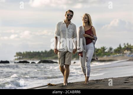 Asie, Indonésie, Bali, jeune couple caucasien marchant le long d'une plage vide et isolée à Bali, tenant les mains, portant des vêtements élégants décontractés, tout en étant sur ho Banque D'Images