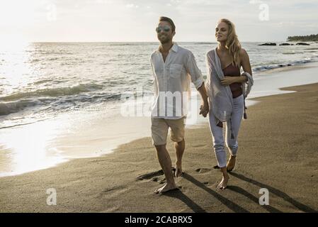 Asie, Indonésie, Bali, jeune couple caucasien marchant le long d'une plage vide et isolée à Bali, tenant les mains, portant des vêtements élégants décontractés, tout en étant sur ho Banque D'Images