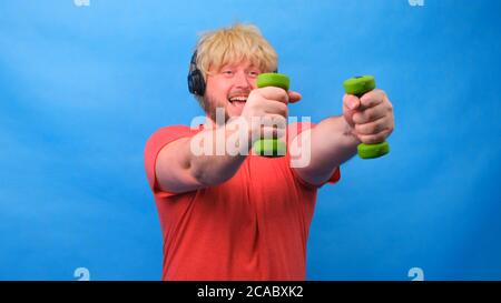 Drôle d'homme de graisse freaky dans une perruque avec des haltères faisant des exercices sur un fond bleu. Banque D'Images