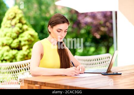 Photo d'une belle jeune femme assise au bureau sur le balcon et écrivant quelque chose. Banque D'Images