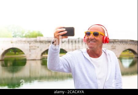 un homme senior prend un selfie et sourit en écoutant de la musique avec des écouteurs - un vieil homme prend un selfie en plein air - concept de retraite heureuse Banque D'Images