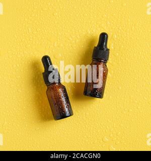 Flacons compte-gouttes en verre ambré avec pipette dans des gouttelettes d'eau sur fond jaune. Flat lay, vue de dessus. Sérum emballage cosmétique, huiles essentielles. Banque D'Images