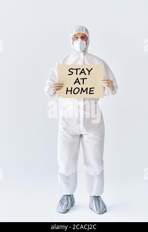 Médecin en costume médical de protection montrant un écriteau vierge rester à la maison sur fond blanc Banque D'Images