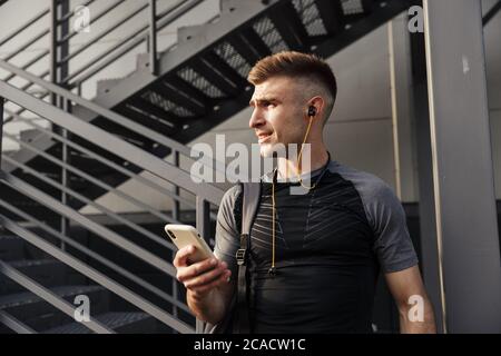 Image d'un jeune sportif heureux avec des écouteurs utilisant un téléphone portable tout en faisant de l'exercice près d'un escalier urbain à l'extérieur Banque D'Images