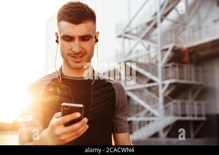 Image d'un jeune sportif satisfait utilisant un téléphone portable et des écouteurs tout en s'exécutant dans un espace urbain extérieur Banque D'Images