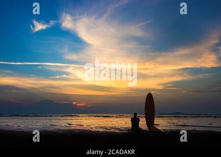 Silhouette d'homme de surf assis avec une planche de surf sur la plage. Scène de surf sur la plage au coucher du soleil avec un ciel coloré. Sports nautiques extérieur aventure style de vie.somme Banque D'Images