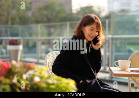 Une belle jeune femme asiatique assise à l'extérieur sous le soleil, faire un appel téléphonique et sourire. Arrière-plan flou Banque D'Images