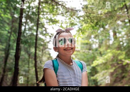 Adorable petite fille portant des lunettes de soleil et souriant tout en appréciant une journée de marche dans une forêt en été Banque D'Images