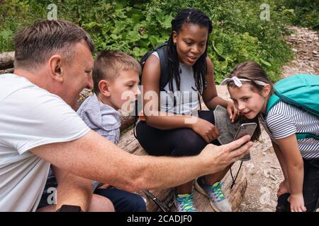 Un père souriant montrant ses deux enfants et un ami des photos sur son téléphone portable lors d'une randonnée ensemble dans une forêt Banque D'Images