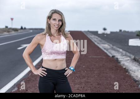 Femme pleine d'assurance, avec les mains à la taille, souriant et regardant l'appareil photo tout en restant debout sur la route pendant l'entraînement par temps nuageux Banque D'Images