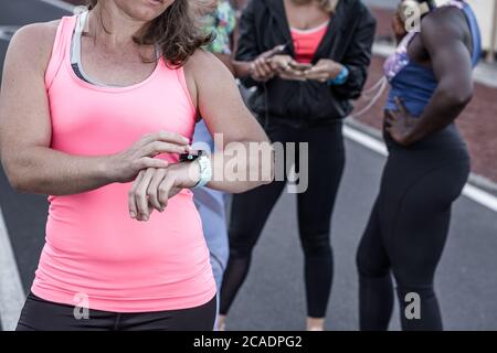 Une athlète féminine forte méconnaissable vérifie le pouls de la montre intelligente tout en restant debout sur la route à proximité de divers amis pendant la séance de course à pied Banque D'Images