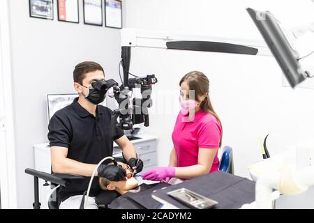 Dentiste dans un uniforme gris et gants en latex regarde dans un microscope dentaire tout en examinant ses dents avec une aide Banque D'Images