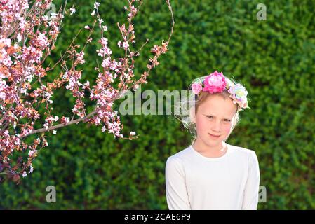 Timide embarrassé belle fille regarde l'appareil photo dans le jardin d'été à la journée ensoleillée.petit enfant mignon avec une couronne de fleur sur sa tête célèbre l'anniversaire ou d'autres vacances.fille de printemps. Banque D'Images