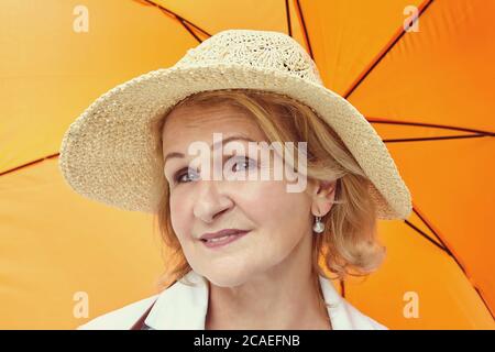 Femme blanche âgée de 62 ans, avec cheveux blonds et chapeau sur sa tête, se tient sous un parapluie orange avec un regard sérieux dans le parc public. Banque D'Images