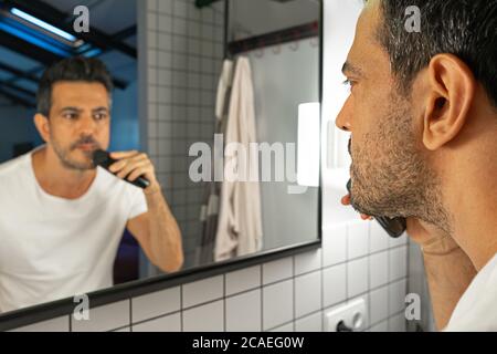 un homme charmant se rassera sa barbe avec une tondeuse à l'avant du miroir de la salle de bains Banque D'Images
