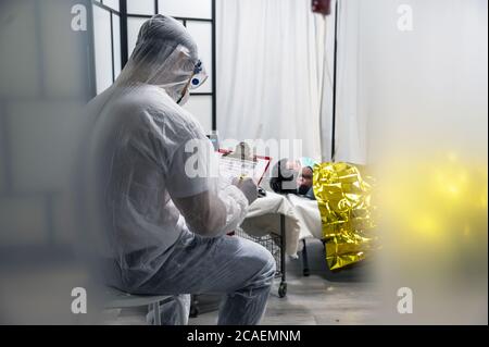 Alerte pandémie Covid-19. Médecin en costume blanc protecteur, lunettes et masque examinent le patient mâle infecté avec une couverture thermique. Banque D'Images