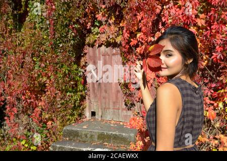 Jeune belle femme asiatique contre la maison de banlieue couverte de feuilles d'automne Banque D'Images