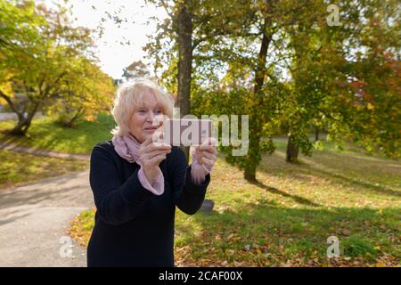 Femme heureuse senior souriant tout en prenant des photos avec un téléphone portable sur le chemin d'un parc naturel calme et reposant Banque D'Images