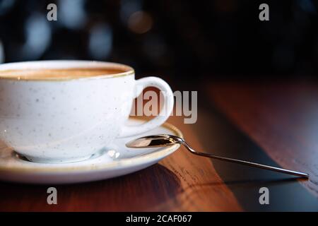 café avec mousse, tasse texturée blanche et cuillère en métal sont sur bâton, gros plan, cappuccino dans la tasse, table en bois brun, céramique, plats en argile, blanc dis Banque D'Images