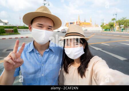 Couple asiatique heureux touristes à voyager porter un masque à protéger de Covid-19 sur eux vacances et selfie par caméra dans le temple Wat Phra Kaew à Bangkok, Banque D'Images