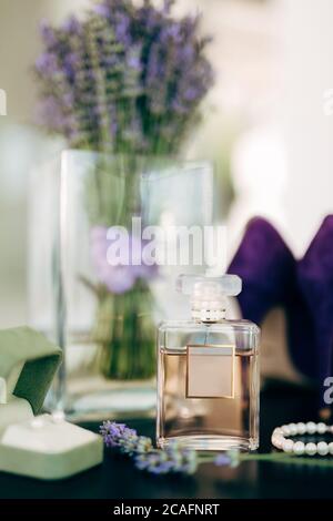 Une bouteille de parfum avec des chaussures lilas pour femmes, un bracelet en perles blanches, une boîte avec des puces d'oreilles et un vase de fleurs sur une surface lisse et sombre. Banque D'Images