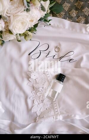 Une bouteille de parfum avec un capuchon noir, un anneau de mariage avec une pierre et un bouquet de mariée de roses blanches sur un tissu blanc avec dentelle avec le mot Bonde. Banque D'Images
