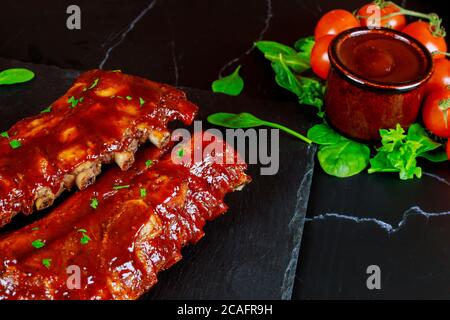 Côtes de porc avec sauce barbecue, tomates et épinards sur panneau en pierre noire. Vue de dessus. Banque D'Images