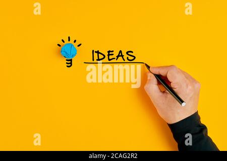 Main masculine écrivant le mot idées à côté d'une ampoule sur fond jaune. Concept d'idées créatives et d'innovation. Banque D'Images