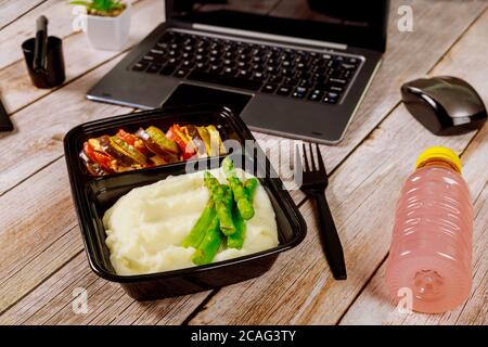 Boîte à lunch contenant une purée de pommes de terre, des asperges et des légumes sur une table en bois avec ordinateur portable. Concept de livraison de nourriture. Banque D'Images