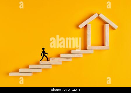 Homme stickman escalade les échelles vers la maison formé par des blocs de bois sur fond jaune. Banque D'Images