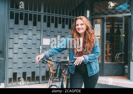 Portrait de mode de sourire rouge courbé cheveux longs caucasienne jeune fille sur la rue de la ville marchant avec le vélo. Les gens naturels beauté de la vie urbaine concept Banque D'Images