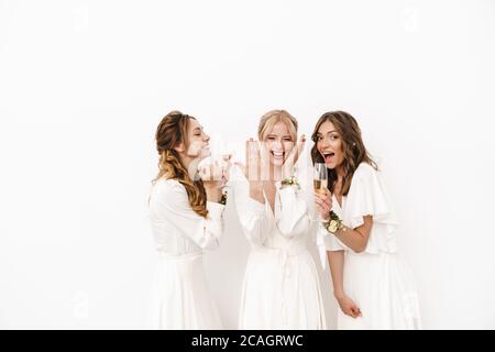 Photo d'une belle mariée heureuse montrant son anneau de mariage à des demoiselles d'honneur excitées isolées sur un mur blanc Banque D'Images